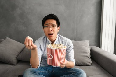 Süre evde bir kanepede oturan ve Tv izlerken hayal kırıklığına uğramış genç Asyalı adam yeme patlamış mısır portresi