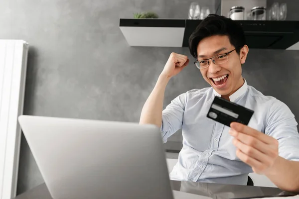 노트북 컴퓨터를 집에서 테이블에 앉아서 플라스틱 카드를 만족된 아시아 남자의 — 스톡 사진