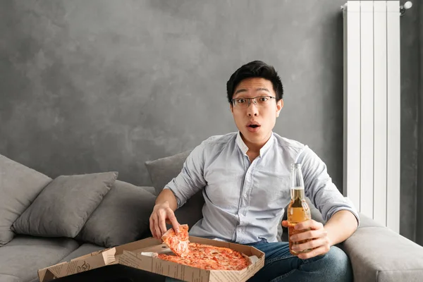 ピザを食べて ビールを飲みながら 自宅でソファに座って テレビを見てショックを受けた若いアジア人の肖像画 — ストック写真