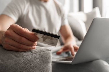 Beyaz adamın 30'lu dizüstü kullanarak ve evde kanepede otururken online alışveriş için kredi kartı tutan rahat kot kırpılmış fotoğraf