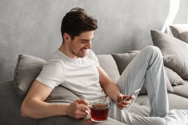Gülümseyen ve cep telefonu evde koltukta çay yudumlarken elinde tutan temel t-shirt yakışıklı adam