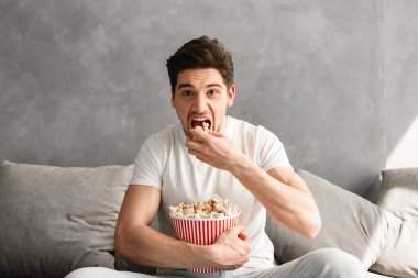 Adam 20'li yaşlarda tek kişilik kanepe gri dairede oturan ve sabah tv izlerken pop mısır yemek