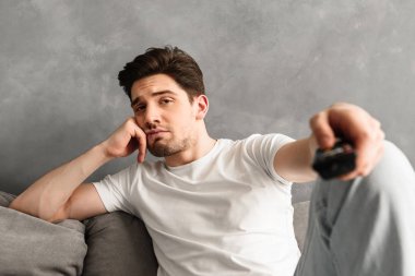 Fotoğraf portre üzgün adam evde kanepede oturan ve uzaktan kumanda kamerada gösteriyor