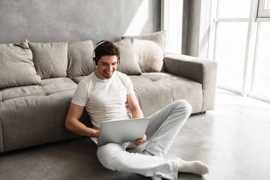 Neşeli adam 30'lu katta evde oturan ve laptop kablosuz kulaklık ile müzik dinlemek temel giyim