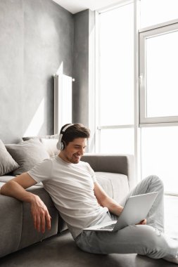 Mutlu adam 30'lu katta evde oturan ve laptop kablosuz kulaklık kullanarak müzik dinlemek temel giyim