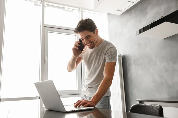 Hombre Morena Optimista Hablando Teléfono Móvil Utilizando Portátil Mesa Cocina — Foto de Stock