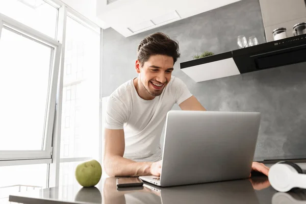 Attractive man 30s in basic clothing looking on laptop while standing in home kitchen