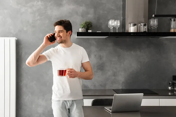 Beautiful cheerful man speaking on mobile phone and looking aside while holding glass with tea