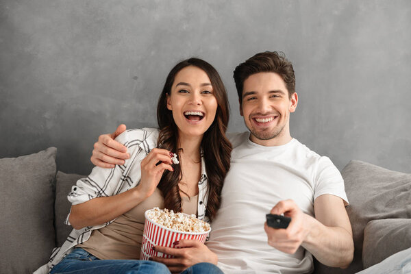 Image of young happy family sitting on sofa at home and looking at you showing remote control at camera and eating pop corn