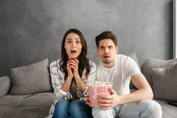 Portrait of tense man and woman sitting on sofa at home and looking at you while watching scary movie and eating popcorn