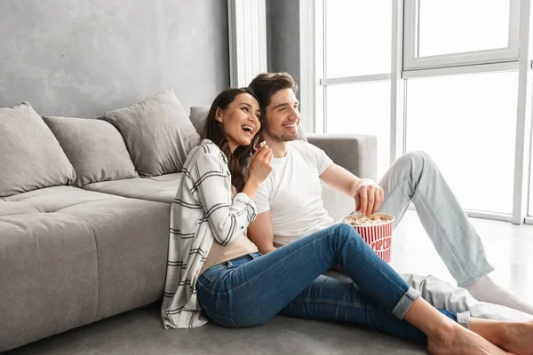 自宅のソファーの近くの床の上に座って ポップコーンを食べながらよそ見笑顔のカップルのイメージ — ストック写真