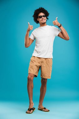 Başparmak yukarıya jest yapmak imge-in neşeli genç Afrika kıvırcık adam kamera arıyorum mavi arka plan üzerinde izole ayakta.