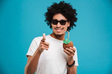 Sana işaret kokteyl Hindistan cevizi içme mavi arka plan üzerinde izole ayakta neşeli Afrika kıvırcık yakışıklı genç görüntü.