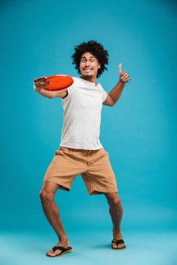 Tutmak beach oyuncak başparmak ile mavi arka plan üzerinde izole ayakta neşeli Afrika kıvırcık yakışıklı genç görüntü.