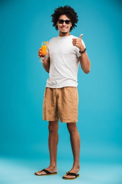Görüntü izole kokteyl gösteren başparmak dayanabiliyor mavi arka plan üzerinde duran Afrika kıvırcık yakışıklı genç.