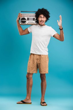 Heyecanlı genç Afrika kıvırcık adam boombox tutan mavi arka plan üzerinde izole ayakta görüntüsünü.