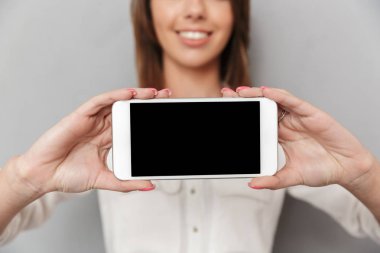 Cep telefonu görüntüsünü gösterilen gri duvar arka plan üzerinde izole ayakta neşeli genç iş kadını görüntü kırpılmış.