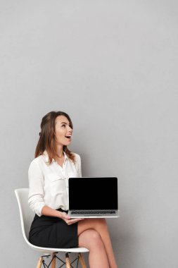 Dizüstü bilgisayarın görüntü gösterilen gri duvar arka plan üzerinde izole görüntüsünü oturan duygusal mutlu genç iş kadını.