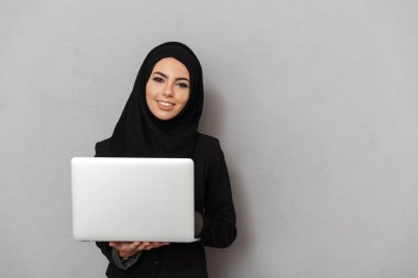 Gri arka plan üzerinde izole dizüstü tutarken kameraya seyir siyah geleneksel Tesettür Arapça gülümseyen kadın 20'li yaşlarda portresi