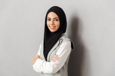 Bir gülümseyen genç Arap kadın katlanmış silah ile ayakta ve gri arka plan üzerinde izole kamera bakarak portre