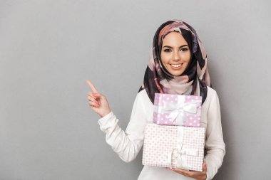 Mevcut kutuları yığını tutan ve uzak gri arka plan üzerinde izole parmak işaret mutlu genç bir Arap kadın portresi