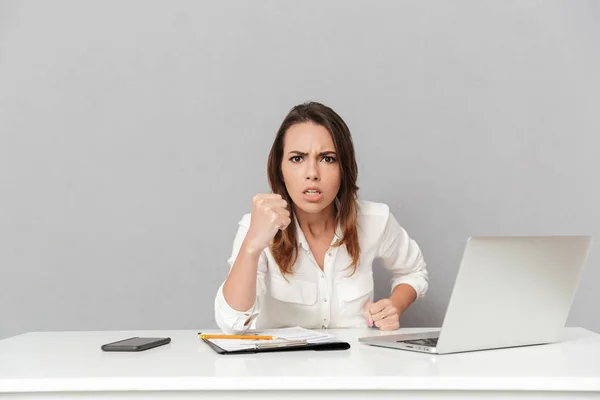 一个愤怒的年轻的商业妇女的肖像坐在办公桌上 并显示她的拳头孤立的白色背景 — 图库照片
