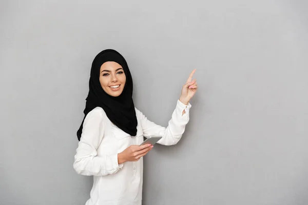 华丽的阿拉伯妇女的肖像在头巾与东方化妆品持有银智能手机和手指在复制空间孤立的灰色背景 — 图库照片