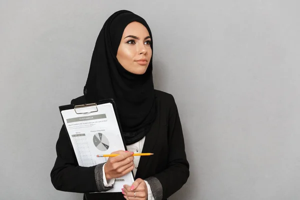 一个快乐的年轻阿拉伯妇女的肖像持有剪贴板与图表 并看着远离灰色背景孤立 — 图库照片