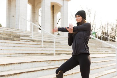 Closeup image of beautiful handicapped sportswoman in tracksuit with prosthesis warming up and stretching at the stairs outside clipart