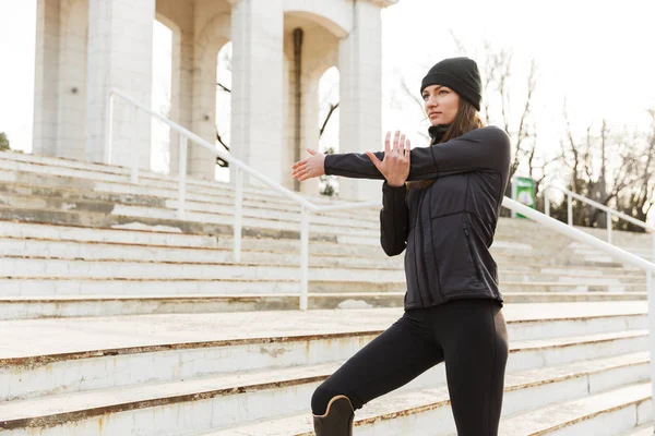Closeup image of beautiful handicapped sportswoman in tracksuit with prosthesis warming up and stretching at the stairs outside