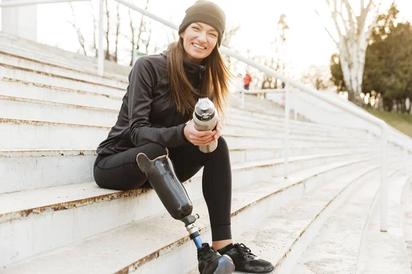 Strong willed handicapped woman in black tracksuit with prosthetic leg laughing while sitting at the street stairs with thermos cup in hands
