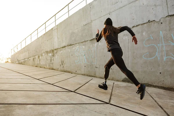 Portrait of disabled athlete woman with prosthetic leg in tracksuit running along concrete wall outside