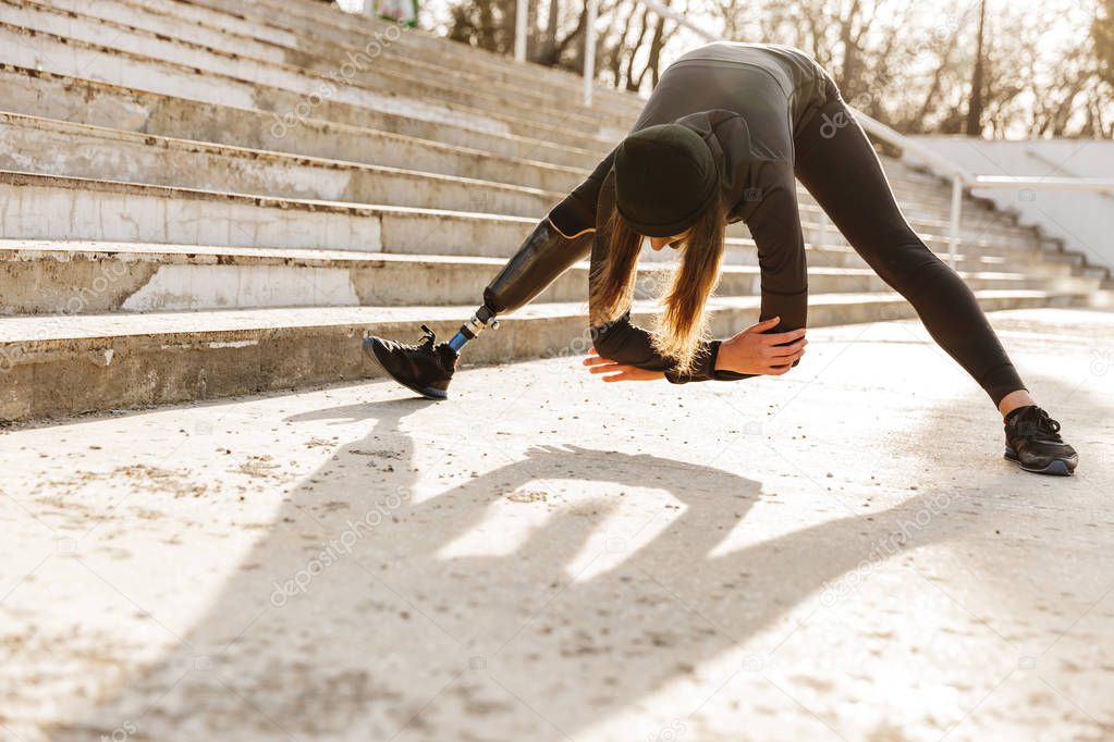 Image of disabled sportswoman in black tracksuit having prosthetic leg exercising with slopes and stretching body outdoors
