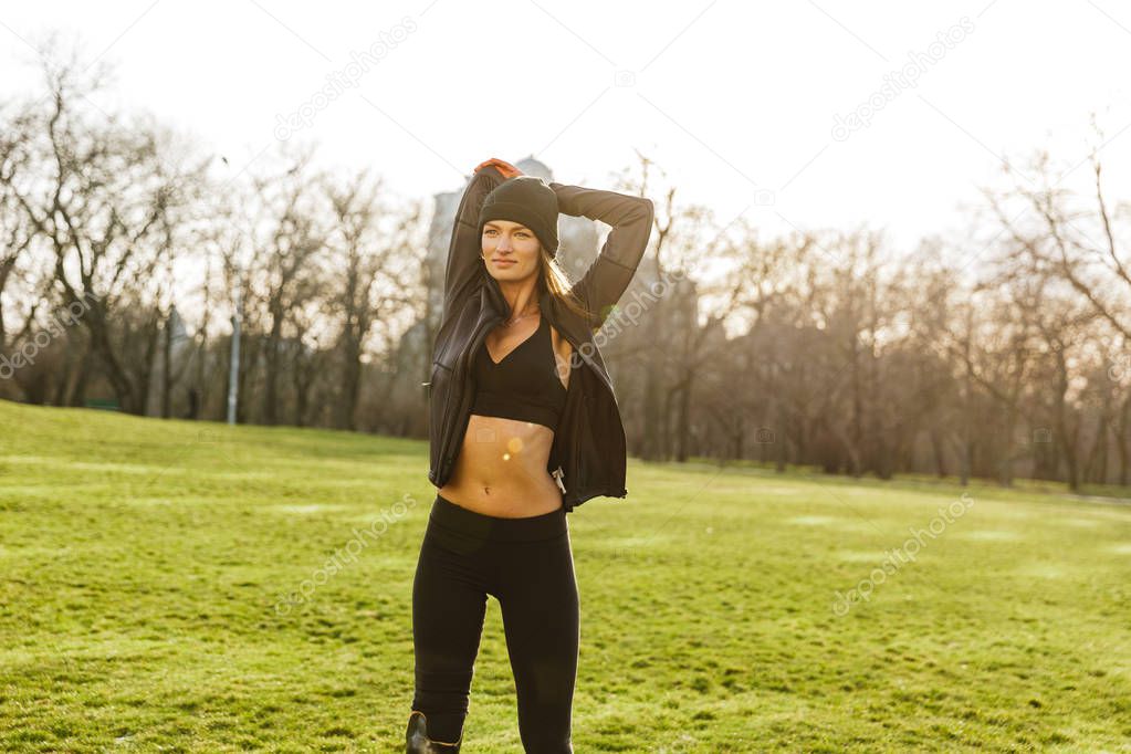 Image of attractive handicapped sportswoman in tracksuit with prosthesis leg warming up on grass and stretching arms
