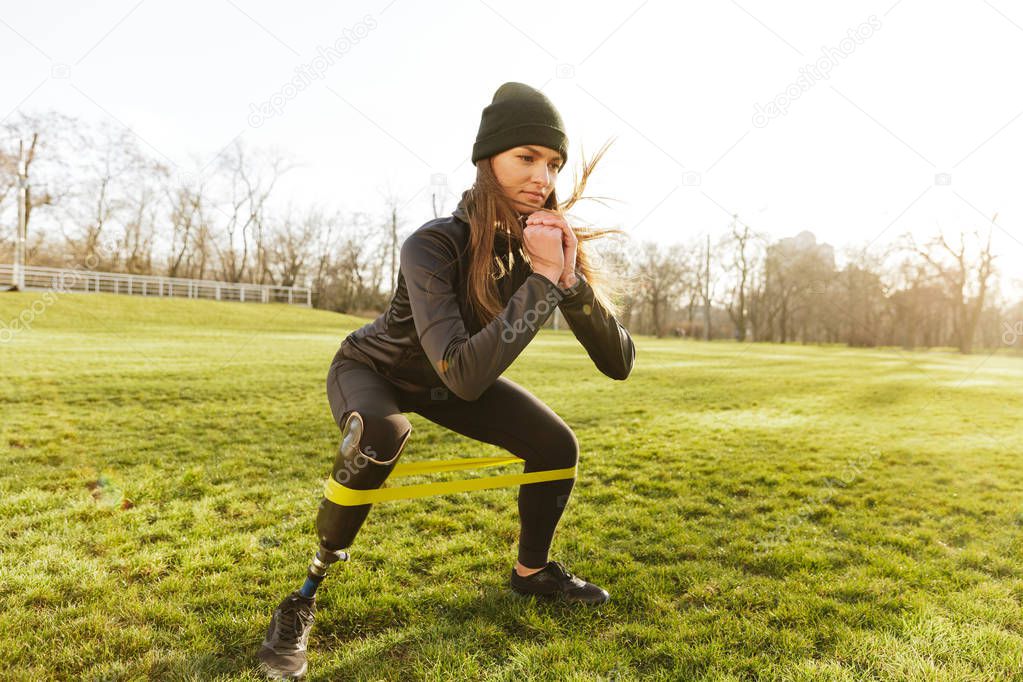 Photo of concentrated disabled girl in tracksuit doing sports and squatting with prosthetic leg on grass using resistance band