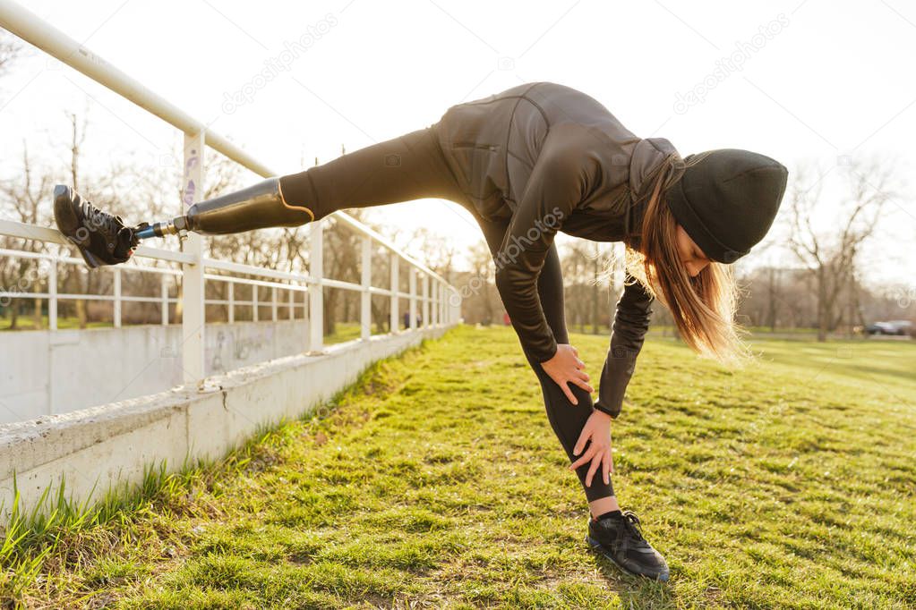 Photo of disabled running woman in sportswear doing slopes and stretching prosthetic leg on grass using railing