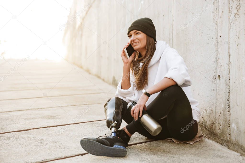 Photo of happy handicapped woman in casual wear having bionic leg sitting on concrete floor outside and talking on mobile phone