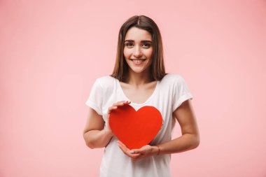 Pembe arka plan üzerinde izole kırmızı kalp tutan mutlu bir genç kadın portresi