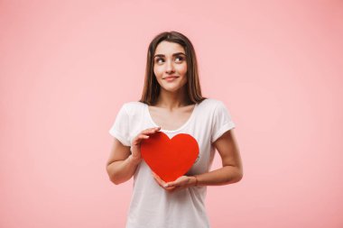 Pembe arka plan üzerinde izole kırmızı kalp tutan bir gülümseyen genç kadın portresi