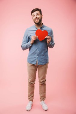 Pembe arka plan üzerinde izole kırmızı kalp tutan mutlu bir genç adamın tam uzunlukta