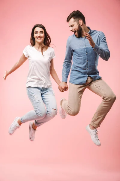 ピンクの背景に分離されたジャンプしながら手を繋いでいる興奮している若いカップルの完全な長さの肖像画 — ストック写真