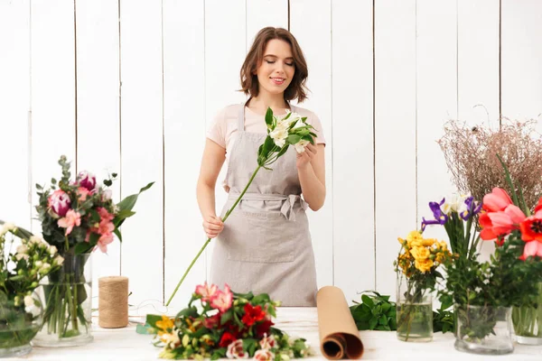 Young pretty smiling florist woman standing near table with different flowers and looking aside in workshop.