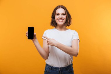 Sarı arka plan görüntüsünü cep telefonu tarafından gösterilen üzerinde izole ayakta heyecanlı genç kadın görüntüsü. Görünümlü kamera.