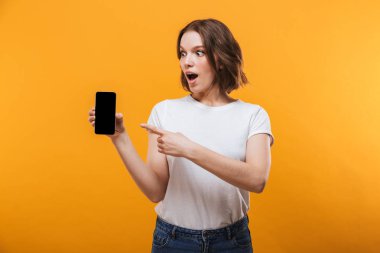 Sarı arka plan görüntüsünü cep telefonu tarafından gösterilen üzerinde izole ayakta heyecanlı genç kadın görüntüsü. Bir kenara arıyorsunuz.