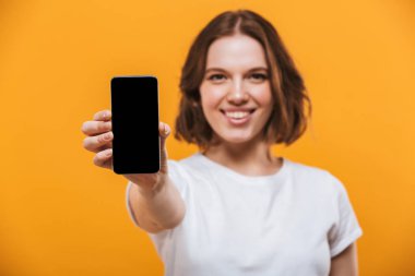 Mutlu genç kadının görüntüsünü cep telefonu tarafından gösterilen sarı arka plan üzerinde izole ayakta fotoğrafını. Görünümlü kamera.