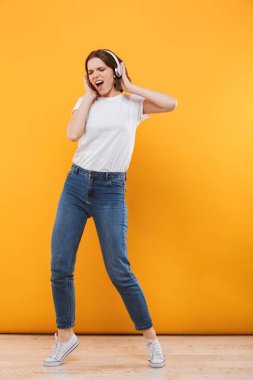 Tam uzunlukta kulaklık ile müzik dinlemek ve sarı arka plan üzerinde dans kazaklı mutlu bir genç kadın portresi