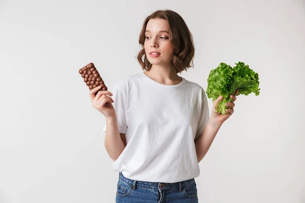 一个困惑的年轻女子的肖像持有巧克力棒和生菜隔离在白色背景 — 图库照片