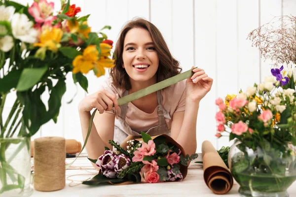 ワーク ショップの花で作業中のテーブルの近くに立ってかわいい陽気な花屋の女性の写真 カメラを探しています — ストック写真