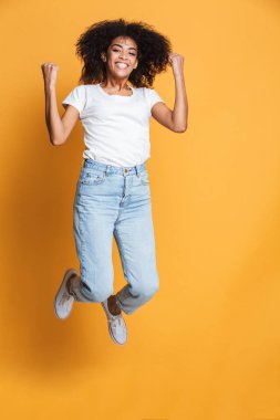 Neşeli bir genç Afrikalı kadın atlama izole sarı arka plan üzerinde iken başarı kutluyor tam uzunlukta portresi