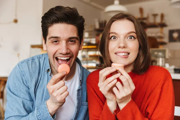 Funny image of beautiful happy couple man and woman looking at camera and eating macaron biscuits